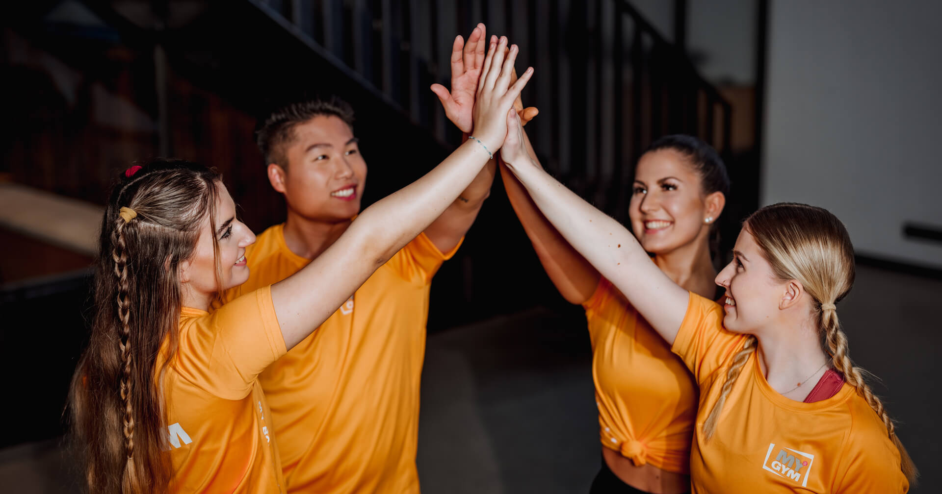 Vier junge Mitarbeiter in orangefarbenen „MY GYM“-T-Shirts stehen im Kreis und geben sich ein triumphales High-Five. Sie lächeln einander motiviert an. Die Szene symbolisiert Teamgeist und erfolgreiche Zusammenarbeit in einem modernen Fitnessstudio-Umfeld.