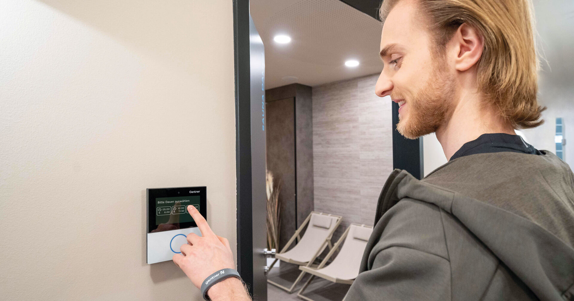 Ein junger Mann mit blonden Haaren und Bart bedient lächelnd ein kleines, an der Wand installiertes Touchscreen-Terminal der Marke Gantner.