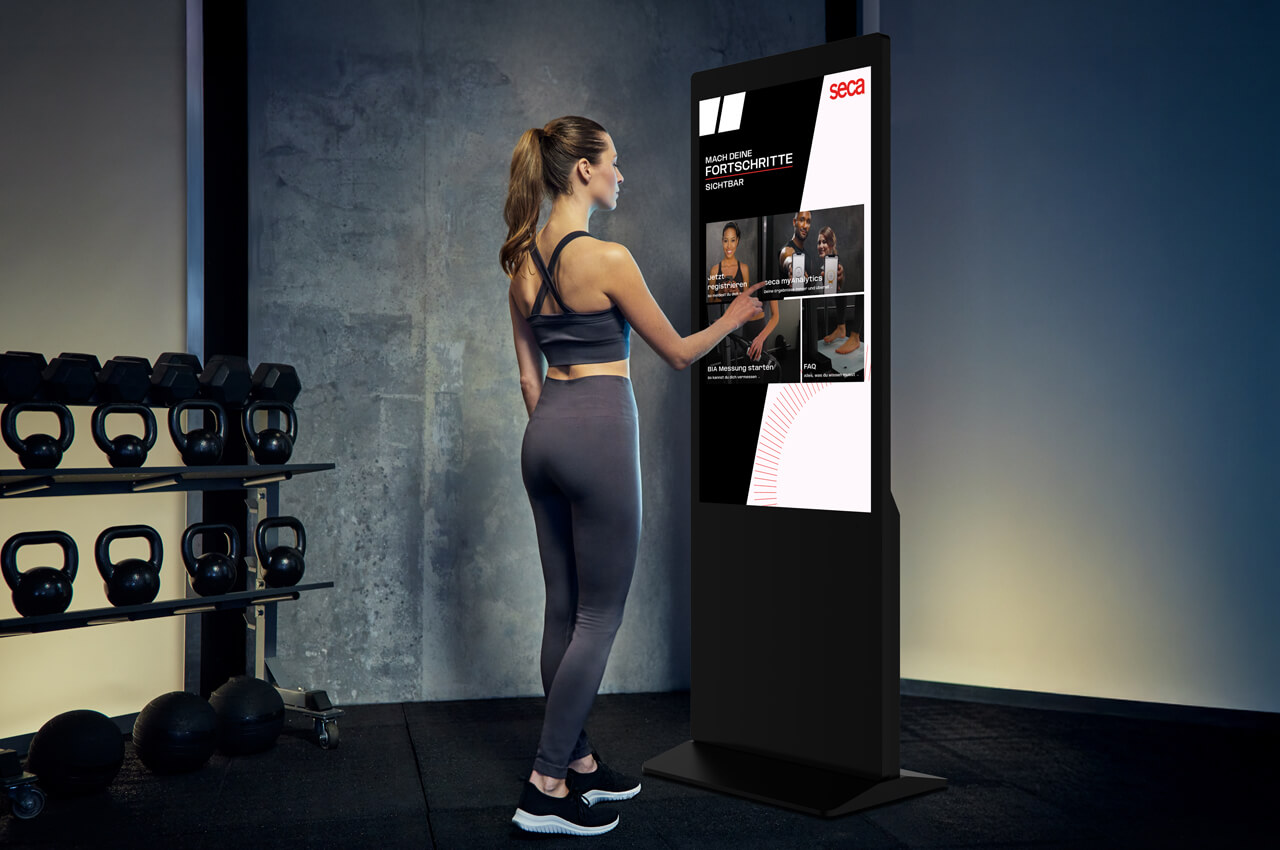 Eine sportliche Frau in grauer Trainingskleidung bedient den großen, aufrechten seca TRU Companion Touchscreen in einem Fitnessstudio. Der Bildschirm zeigt Onboarding-Inhalte, Videotutorials und die Aufforderung: "MACH DEINE FORTSCHRITTE SICHTBAR".
