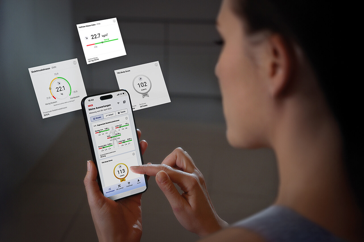 Eine Person hält ein Smartphone, das die seca Cloud Mitglieder-App mit der Ansicht "Meine Auswertungen" zeigt. Das Display präsentiert verschiedene Gesundheits- und Fitness-Scores (z.B. TBW Body Score) und Diagramme, die die Trainingsfortschritte visualisieren.