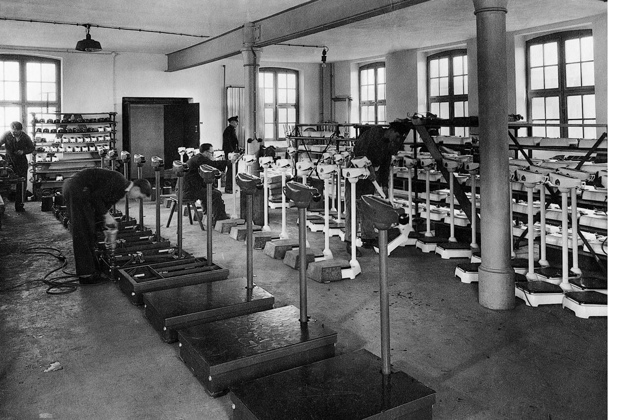 Schwarz-Weiß-Aufnahme aus einer historischen seca-Fabrik- oder Montagehalle. Mehrere Mitarbeiter in Arbeitskleidung montieren und kalibrieren in Reihen aufgestellte, klassische Säulenwaagen (Personenwaagen) in einem lichtdurchfluteten Raum mit hohen Fenstern und Stahlträgern.