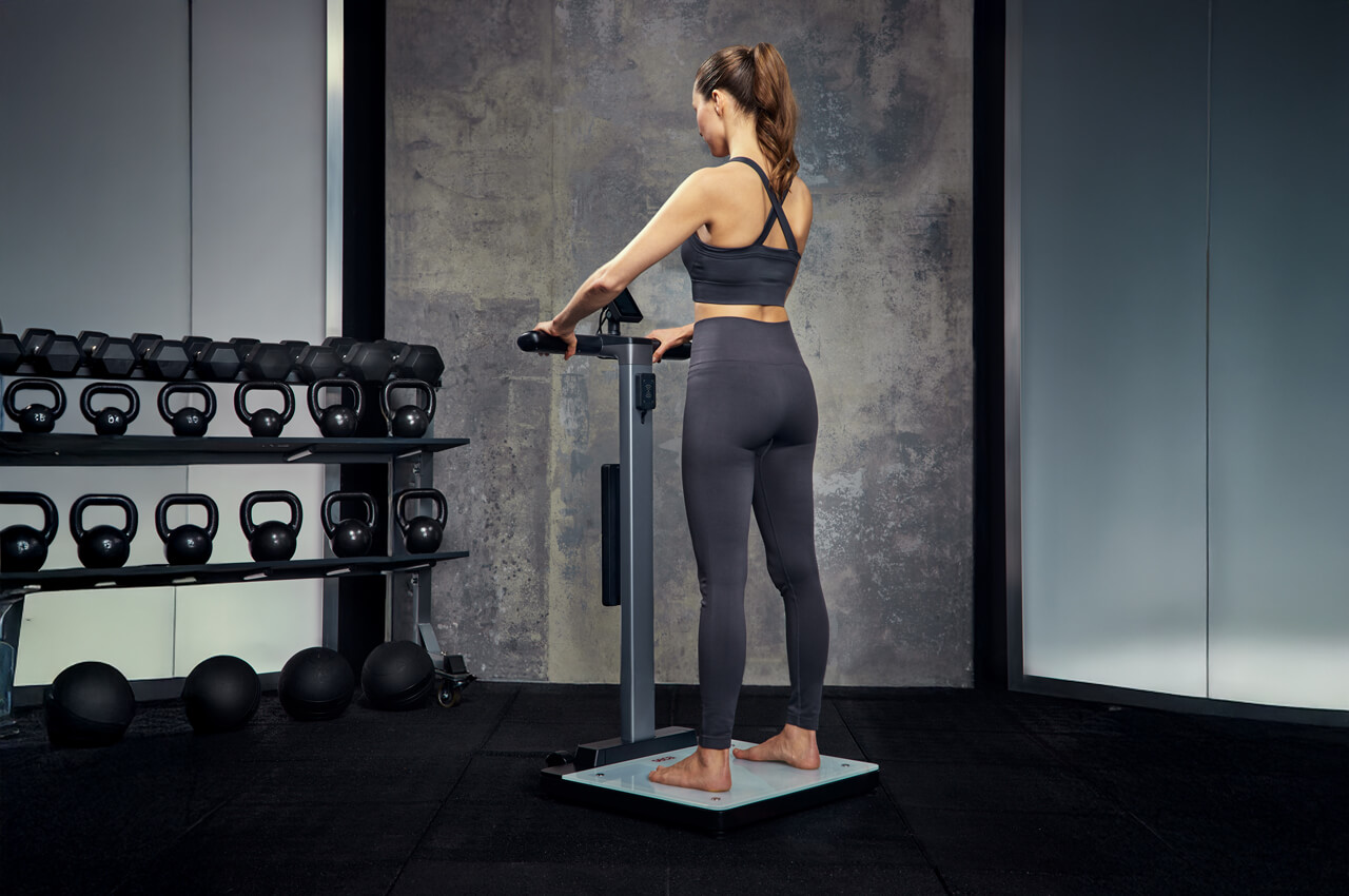 Seitenansicht einer Frau, die barfuß auf der weißen Standfläche der seca TRU steht und sich an den Handelektroden (Griffen) festhält, um eine Bioimpedanz-Messung durchzuführen. Im Hintergrund steht ein Regal mit Kettlebells.