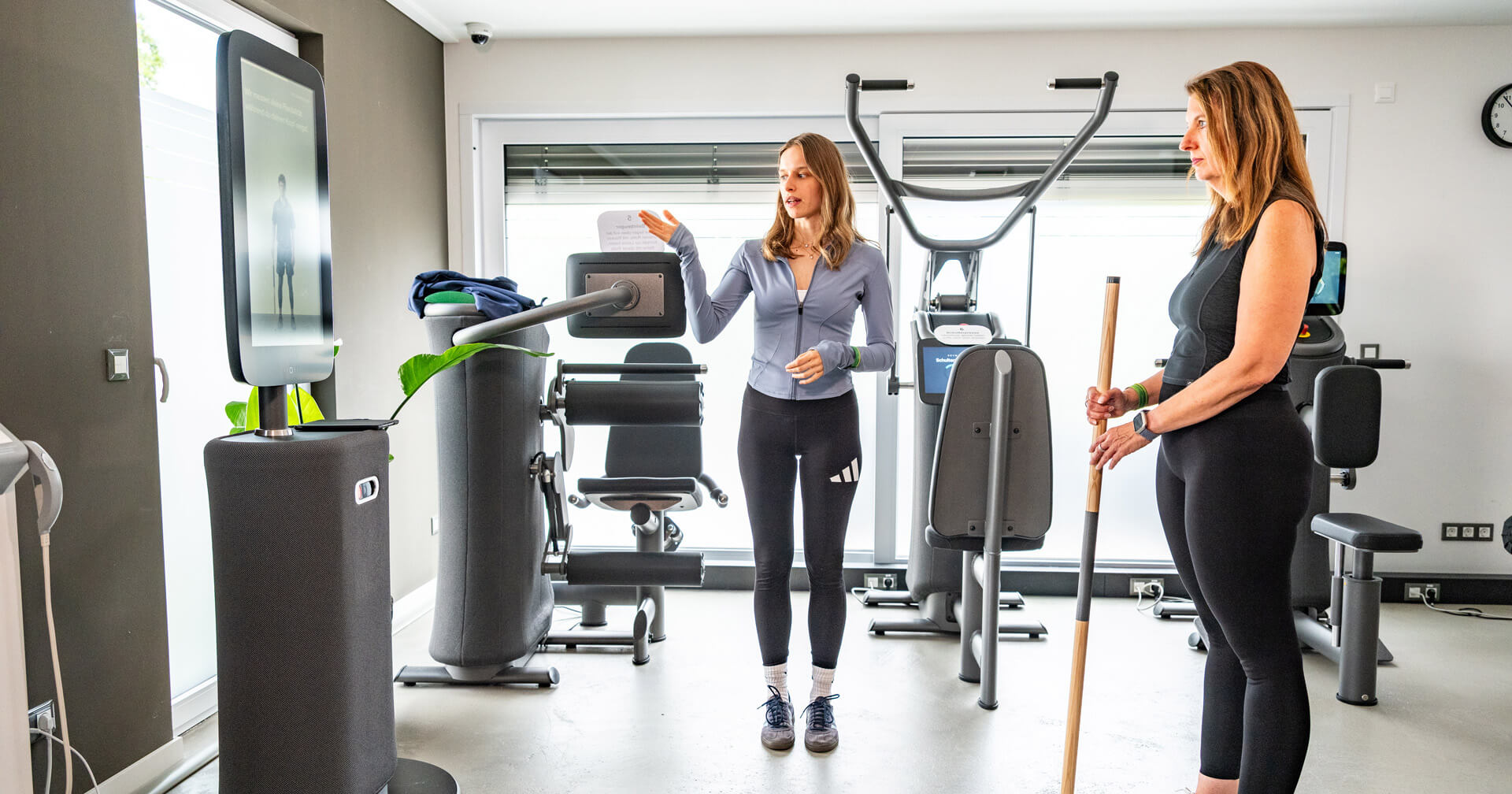 Eine junge Trainerin in sportlicher Kleidung erklärt einer Patientin die Nutzung eines digitalen Trainingssystems in einer modern ausgestatteten Physiotherapiepraxis. Die Trainerin steht in der Bildmitte und gestikuliert in Richtung eines großen Terminal-Bildschirms (EGYM Hub) auf der linken Seite.