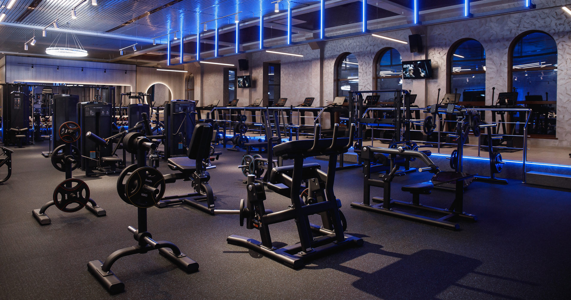 Ein großer, modern gestalteter Trainingsbereich eines Fitnessstudios bei gedimmter, blauer LED-Deckenbeleuchtung. Im Vordergrund stehen verschiedene schwarze Krafttrainingsgeräte, Hantelständer und Gewichtsscheiben auf einem dunklen Gummiboden.