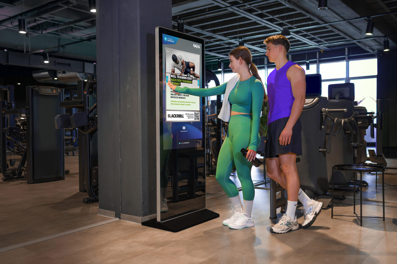 Zwei Personen in Sportkleidung stehen in einem modern ausgestatteten Fitnessstudio vor einem gro&szlig;en, freistehenden Touchscreen-Terminal. Eine Frau in einem gr&uuml;nen Sport-Set bedient den Bildschirm mit der Hand, w&auml;hrend ein Mann in einem lila Muskelshirt daneben steht und zuschaut. Auf dem Display ist eine Werbe- oder Trainingsanzeige zu sehen.