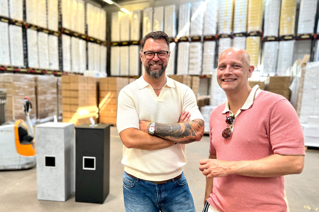 Zwei l&auml;chelnde M&auml;nner stehen nebeneinander in einer gro&szlig;en Lagerhalle, die bis oben hin mit Kartons und Paletten gef&uuml;llt ist. Links im Hintergrund sind zwei Hygiene-Spenders&auml;ulen in Beton-Optik und Schwarz zu sehen.