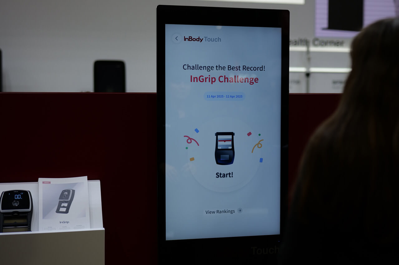 Nahaufnahme eines &bdquo;InBody Touch&ldquo;-Bildschirms mit der Anzeige &bdquo;InGrip Challenge &ndash; Challenge the Best Record!&ldquo;. Auf dem Display ist eine spielerische Grafik mit Konfetti zu sehen. Im Vordergrund links steht ein gedruckter Prospekt des InGrip-Handkraftmessger&auml;ts, rechts ist die Silhouette einer Person von hinten zu erkennen.
