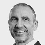  Schwarz-Weiß-Porträt eines lächelnden Mannes (Uwe Schoch) mittleren Alters mit kurzem Bartansatz und Anzug vor hellem Hintergrund.