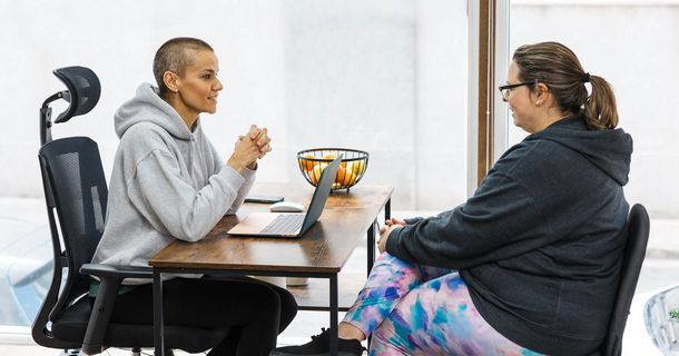 Eine korpulente Frau mit Brille im grauen Hoodie sitzt einer Beraterin mit kurzen Haaren im grauen Hoodie gegenüber. Sie sprechen an einem Holztisch, auf dem ein Laptop und ein Obstkorb stehen. Beide wirken konzentriert und entspannt.