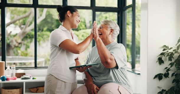 Eine Physiotherapeutin im weißen Polo-Shirt hält ein Tablet im Arm und gibt einer Patientin im hellblauen Shirt ein fröhliches High Five. Beide lächeln herzlich vor einem großen Sprossenfenster einer hellen Praxisraum-Kulisse.