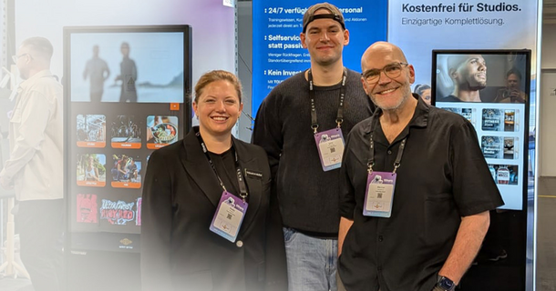 Drei Personen des TOUCH:BAY-Teams stehen lächelnd mit Messeausweisen vor einem Stand der fithera 2026. Im Hintergrund sind Werbebanner mit der Aufschrift „Kostenfrei für Studios“ sowie zwei hochformatige Touchscreen-Displays mit der TOUCH:BAY-Software zu sehen.