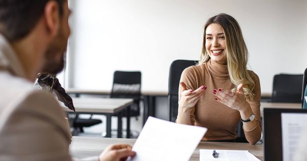 Eine lächelnde junge Frau mit blonden Haaren im braunen Rollkragenpullover sitzt in einem modernen Büro an einem Tisch und gestikuliert lebhaft während eines Gesprächs. Ihr Gegenüber ist nur von hinten im Anschnitt zu sehen und hält ein Dokument in der Hand.