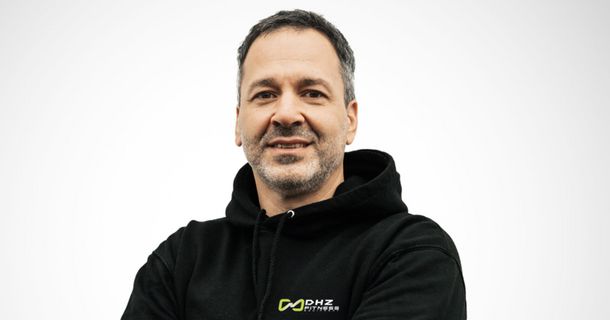 Porträt des DHZ Fitness Business Solutions Key Account Parwis Hafez-Ghorani, der lächelnd in einem schwarzen Kapuzenpullover mit dem DHZ FITNESS Logo vor einem hellen Hintergrund posiert.