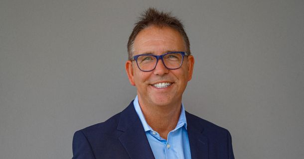 Porträt eines lächelnden Mannes mit kurz geschnittenem, leicht grau meliertem Haar. Er trägt eine markante, blau umrandete Brille, ein blaues Hemd und einen dunklen Blazer. Der Hintergrund ist ein schlichter, neutraler Grauton.