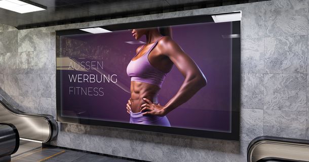In einem Bahnhof hängt ein großformatiges Plakat mit einer sportlichen Frau und dem Schriftzug "Aussen Werbung Fitness"
