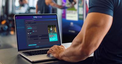 Ein Mann mit muskulösem Arm bedient einen Laptop im Fitnessstudio. Auf dem Bildschirm ist die deutsche Benutzeroberfläche von „my airtango“ zu sehen, auf der gerade unter dem Titel „NEUE NAVIGATOR APP ERSTELLEN“ ein Werbebild mit dem Slogan „BLEIB AUF DEM NEUESTEN STAND!“ bearbeitet wird. Der Hintergrund zeigt eine verschwommene Gym-Umgebung.