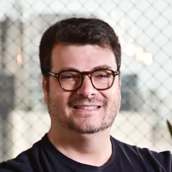 Aggregatoren im Check: Wellhub & EGYM Wellpass beziehen Stellung - 2 Porträt eines lächelnden Mannes mit dunklen Haaren, Bartstoppeln und einer markanten Brille mit dunklem Rahmen. Er trägt ein schwarzes T-Shirt und steht vor einem hellen, strukturierten Hintergrund, der an ein feines Gitter oder eine Glasfassade erinnert.