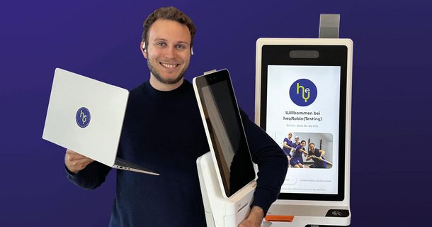heyRobin-Co-Founder Robin Louis Schmidt präsentiert Hardware-Komponenten wie Laptop, Terminal und Check-in-Kiosk für den Praxisempfang.