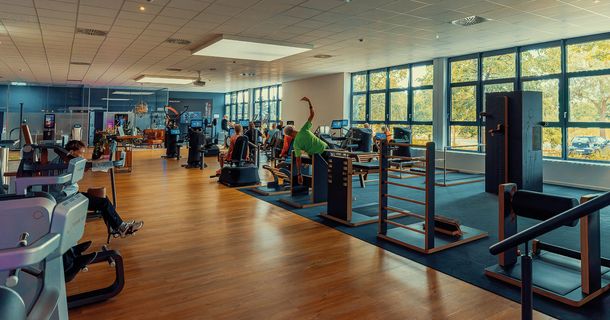 Blick über die große, lichtdurchflutete Trainingsfläche im next level Therapie- und Trainingszentrum in Schweinfurt. Links stehen moderne Kraft- und Ausdauergeräte auf Holzfußboden, während rechts auf dunklem Belag ein Holz-Beweglichkeitszirkel und Sprossenwände vor einer langen Fensterfront angeordnet sind.