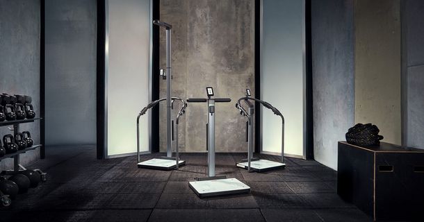 Drei medizinische Mess- und Analysegeräte von seca stehen zentral auf einem dunklen Gummiboden in einem modern gestalteten Fitnessstudio mit Sichtbetonwänden.