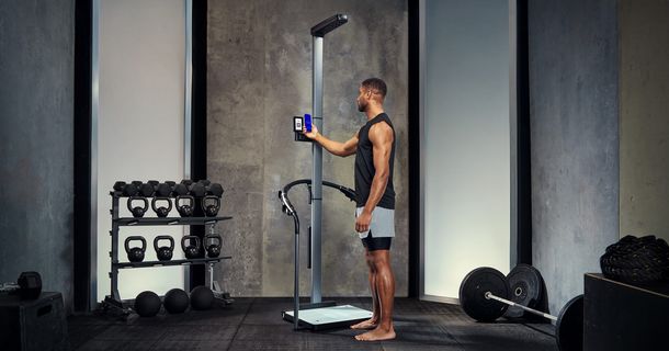 Ein sportlicher, barfüßiger Mann in schwarzem Tanktop und kurzer Sporthose steht auf einer Körperanalysewaage oder einem medizinischen Analysegerät in einem modernen Fitnessstudio. Er bedient das Gerät über einen Touchscreen, der an einer hohen Metallsäule befestigt ist. Im Hintergrund befinden sich eine Hantelbank mit Kettlebells und Hanteln, eine Langhantel mit Gewichten sowie eine Wand mit Betonoptik.