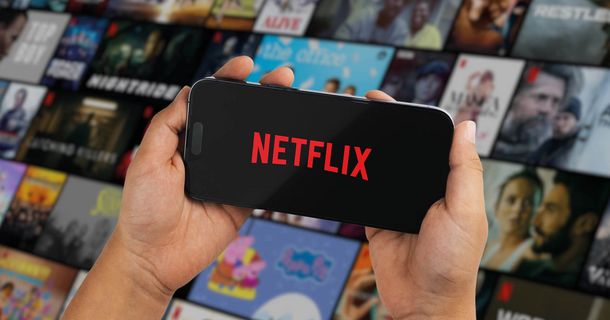 Nahaufnahme von zwei Händen, die ein Smartphone waagerecht halten. Auf dem schwarzen Bildschirm leuchtet das rote Netflix-Logo. Der Hintergrund zeigt eine unscharfe Galerie mit zahlreichen Film- und Serien-Vorschaubildern.