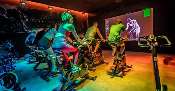 In einer DarkCycling Class sind mehrere Sportler auf Indoor Bikes zu sehen, die auf einen Bildschirm blicken.
