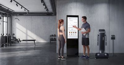 Ein Trainer mit Klemmbrett erklärt einer Sportlerin in einem modern gestalteten Fitnessstudio mit Sichtbetonwänden die Auswertungen auf einem „InBody Touch“-Terminal. Rechts daneben stehen eine professionelle InBody-Körperanalysewaage und ein InGrip-Messgerät auf einem Ständer.