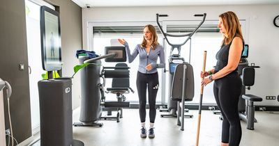 Eine junge Trainerin in sportlicher Kleidung erklärt einer Patientin die Nutzung eines digitalen Trainingssystems in einer modern ausgestatteten Physiotherapiepraxis. Die Trainerin steht in der Bildmitte und gestikuliert in Richtung eines großen Terminal-Bildschirms (EGYM Hub) auf der linken Seite.