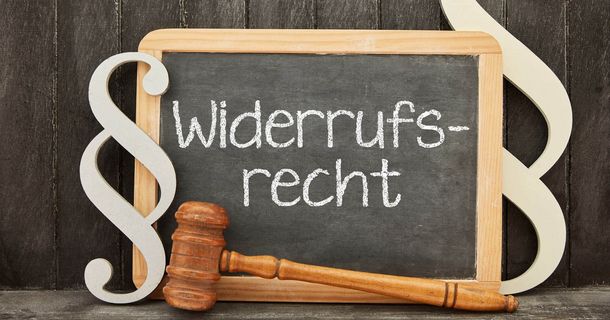 Eine kleine Schiefertafel in einem Holzrahmen, auf der mit Kreide „Widerrufs-recht“ geschrieben steht. Davor liegt ein hölzerner Richterhammer, und links daneben ist ein großes Paragrafenzeichen-Symbol aufgestellt. Der Hintergrund besteht aus dunklen Holzplanken.