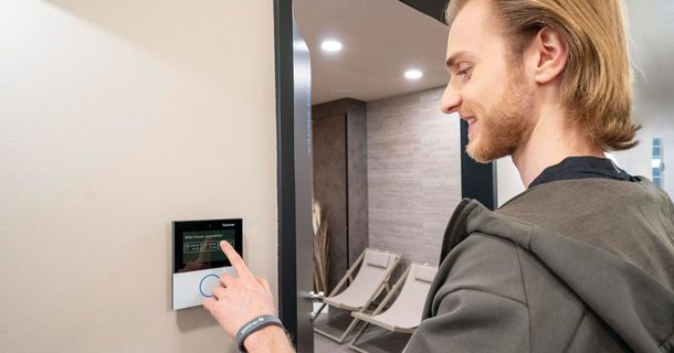 Ein junger Mann mit blonden Haaren und Bart bedient lächelnd ein kleines, an der Wand installiertes Touchscreen-Terminal der Marke Gantner.