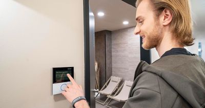 Ein junger Mann mit blonden Haaren und Bart bedient lächelnd ein kleines, an der Wand installiertes Touchscreen-Terminal der Marke Gantner.
