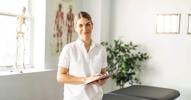 New Pay – Neue Gehaltsstrategie in der therapeutischen Praxis Eine lächelnde, blonde Therapeutin oder Praxis-Mitarbeiterin in weißer Arbeitskleidung (Polohemd) blickt direkt in die Kamera, während sie ein Klemmbrett hält. Sie steht in einem hellen Behandlungs- oder Untersuchungsraum mit einer dunklen Behandlungsliege. Im Hintergrund sind medizinische Wandbilder (Anatomie-Poster) und ein menschliches Skelettmodell neben einem Fenster zu sehen.