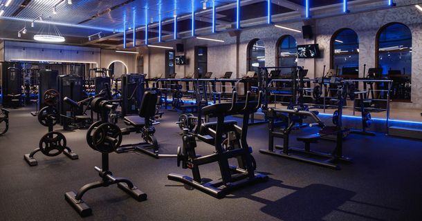Ein großer, modern gestalteter Trainingsbereich eines Fitnessstudios bei gedimmter, blauer LED-Deckenbeleuchtung. Im Vordergrund stehen verschiedene schwarze Krafttrainingsgeräte, Hantelständer und Gewichtsscheiben auf einem dunklen Gummiboden.