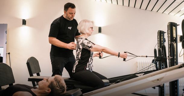 Ein Physiotherapeut im schwarzen Shirt korrigiert die Haltung einer Frau, die auf der Bank eines KGG-Zuggeräts sitzt und eine Zugübung ausführt. Im Vordergrund ist eine weitere Person auf einer Trainingsbank sichtbar.