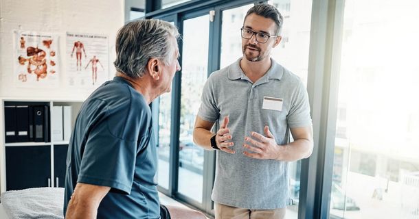 Physiotherapeut erklärt einem im Behandlungsraum sitzenden älteren Patienten gestikulierend den Therapieplan.