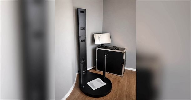 Ein 3D-Körperanalyse-System steht in der Ecke eines Raumes mit grauen Wänden und hellem Holzboden. Das System besteht aus einer runden, schwarzen Bodenplattform mit einer integrierten Waage (Trittfläche mit Fußsymbolen), zwei Haltegriffen und einer hohen, vertikalen Sensorsäule mit mehreren Kameras. Daneben steht ein stabiles, schwarzes Transportcase