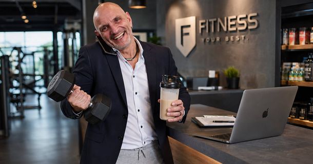 Mann mit Glatze und im Sakko steht mit einer hantel und einem Shake an der Theke in einem Fitnessstudio