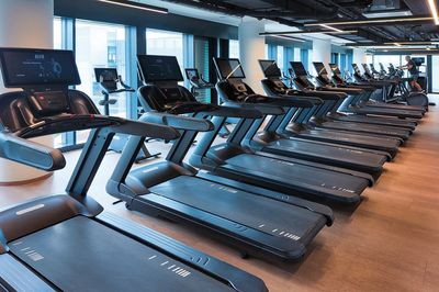 Reihe moderner Laufbänder mit integrierten Bildschirmen in einem hellen Fitnessstudio mit großen Fenstern.