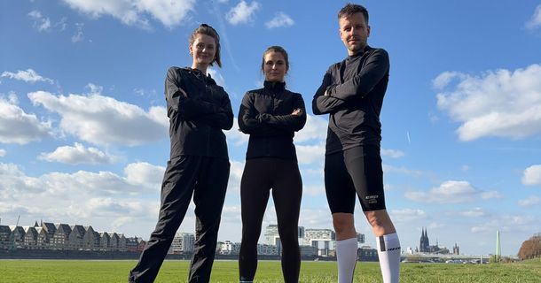 Zwei Frauen und ein Mann im Läufer-Outfit auf einer Wiese unter blauem Himmel