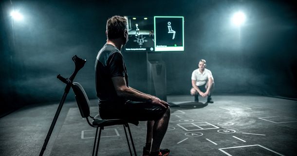 Ein Patient sitzt im Profil auf einem Stuhl, neben ihm lehnt eine Gehstütze (Krücke). Er blickt auf einen Bildschirm des SKILLCOURT-Systems, der eine Bewegungsanalyse und Übungsanleitung anzeigt. Im hintergrundnahen Bereich kniet ein Therapeut und beobachtet die Szene.