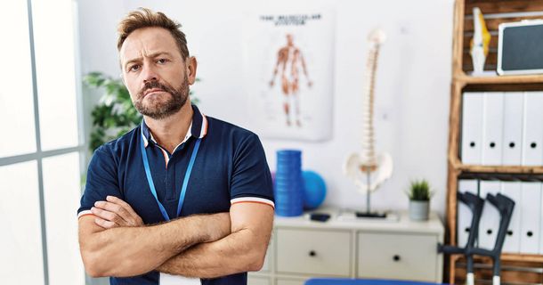 Physiotherapeut blickt mit verschränkten Armen und nachdenklichem, kritischem Blick direkt in die Kamera.