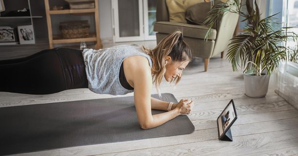 Eine Frau trainiert in ihrem Wohnzimmer auf einer Fitnessmatte und hält die Unterarmstütz-Position (Plank). Sie schaut dabei auf ein Tablet, das vor ihr auf dem Boden steht und ein Workout-Video zeigt. Die Umgebung ist hell und mit Pflanzen und einem Sessel wohnlich gestaltet.