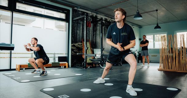 Zwei sportliche Männer stehen auf schwarzen, mit weißen Kreisen markierten AIRIS-Trainingsmatten am Boden. Der Mann im Vordergrund führt einen seitlichen Ausfallschritt aus, während der Mann dahinter in die Kniebeuge geht. Im Hintergrund beobachtet ein Therapeut im schwarzen Shirt die Übungsausführung.