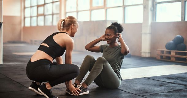 Junge, fitte dunkelhäutige Frau lacht, während sie mit einer blonden Frau im Fitnessstudio Sit-ups macht.