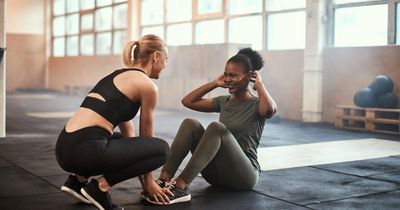 Junge, fitte dunkelhäutige Frau lacht, während sie mit einer blonden Frau im Fitnessstudio Sit-ups macht.