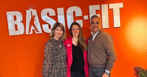 Anke Rekos, Maike Kumstel und Pierre Coolen stehen vor dem BASIC-FIT Logo in der BASIC-FIT-Deutschland-Zentrale