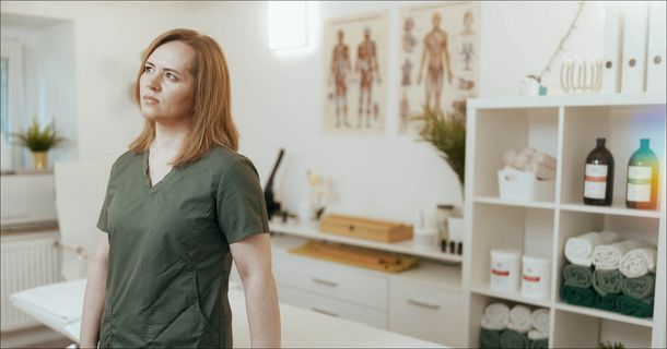 Eine Physiotherapeutin in dunkelgrüner Arbeitskleidung steht mit nachdenklichem, leicht besorgtem Blick in einer Physiotherapiepraxis und schaut zur Seite. Im hellen, leicht unscharfen Hintergrund ist die Praxiseinrichtung zu sehen, darunter eine Behandlungsliege, anatomische Poster an der Wand sowie ein weißes Regal mit Handtüchern und Flaschen.