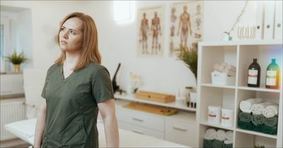 Eine Physiotherapeutin in dunkelgrüner Arbeitskleidung steht mit nachdenklichem, leicht besorgtem Blick in einer Physiotherapiepraxis und schaut zur Seite. Im hellen, leicht unscharfen Hintergrund ist die Praxiseinrichtung zu sehen, darunter eine Behandlungsliege, anatomische Poster an der Wand sowie ein weißes Regal mit Handtüchern und Flaschen.