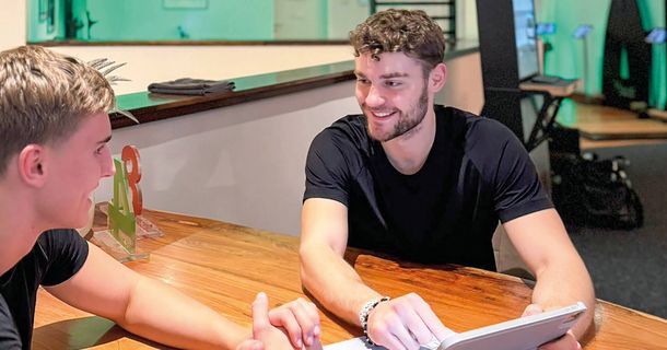 Zwei junge Männer in schwarzer Sportkleidung sitzen sich an einem rustikalen Holztisch gegenüber. Einer der Männer hält ein Tablet in der Hand und erklärt lächelnd Inhalte auf dem Bildschirm. Im Hintergrund ist unscharf ein Trainingsbereich mit Fitnessgeräten und grünlicher Beleuchtung zu sehen.